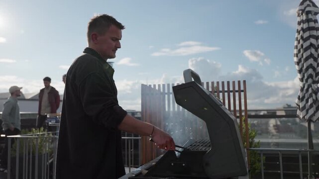 Early evening shot of a man cooking food on a rooftop barbecue in Hamburg, Germany, with friends in background, ideal for lifestyle, leisure, summer, food and social gathering themes