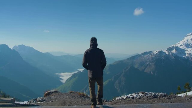 Man hiker snowy mountain valley back view, static tripod shot. Hooded traveler standing and looking at the winter alpine landscape from above.