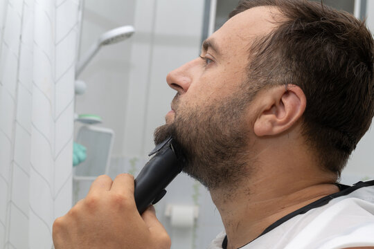 A young man is carefully trimming his beard with an electric trimmer while standing in a modern bathroom. The reflection in the mirror emphasizes his grooming routine.