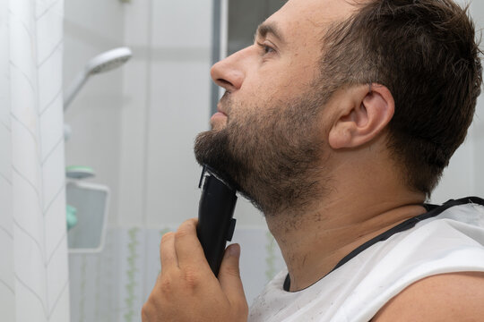 A young man is meticulously trimming his beard in a bathroom, using an electric trimmer for precise grooming. He is wearing a white protective cape.