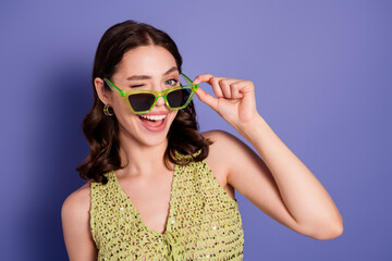 Obraz premium Young woman winking and smiling while adjusting green sunglasses in a playful summer fashion portrait on a purple background