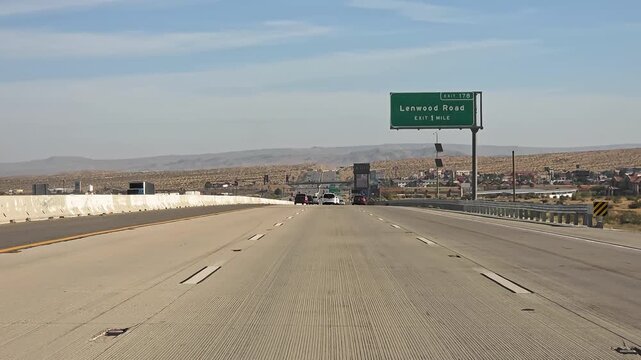 United States March 27 2026, Cars travel on a desert highway toward Lenwood Road exit near Barstow, Wide road video frame shows traffic signs dry hills and open Mojave landscape