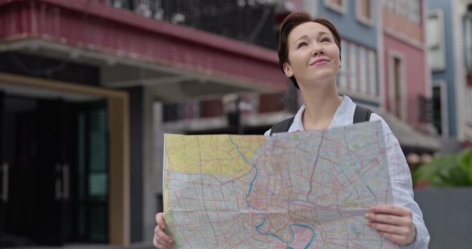 Portrait of happy female tourist holds map and looks around near old buildings. Woman with rucksack walks enjoying cityscape of historical center on vacation
