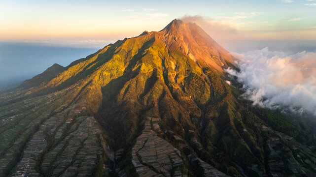 Aerial view of the smoking summit of Mount Merapi volcano with terraced agricultural fields and clouds during golden hour in Indonesia.