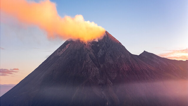 Aerial view of an active volcano with a thick plume of smoke rising from the crater during golden hour with mist at the base in Indonesia.