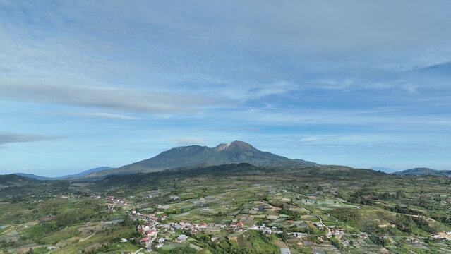 Aerial view of the Alahan Panjang area featuring lush green agricultural fields and a majestic mountain under a clear blue sky in Lembah Gumanti, West Sumatra, Indonesia.
