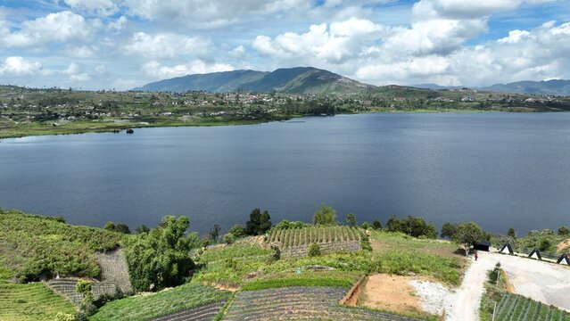 Aerial view of Alahan Panjang featuring the calm lake waters, terraced agricultural fields, and distant mountains under a cloudy sky in Lembah Gumanti, West Sumatra, Indonesia.