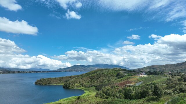 Aerial view of Alahan Panjang featuring a vast blue lake surrounded by lush green hills and a bright cloudy sky in Lembah Gumanti, West Sumatra, Indonesia.