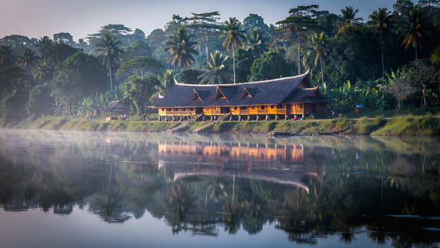 Rumah Betang, a traditional Dayak longhouse, on the banks of a tranquil river in the Borneo rainforest. Perfect reflections in the water, lush tropical vegetation, and morning mist.