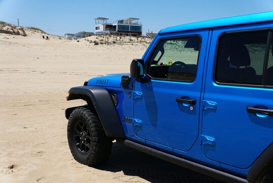 Fenwick Island, Delaware, U.S.A - May 22, 2026 - Bright blue Jeep Wrangler Willy's parked on sandy beach near dunes and modern coastal homes under clear sky.