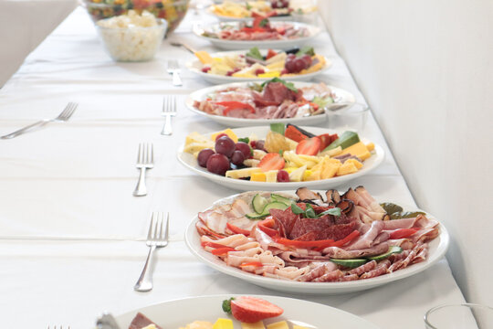 Catering buffet line with plates of assorted cold cuts, meats and cheese platters on a white tablecloth.
