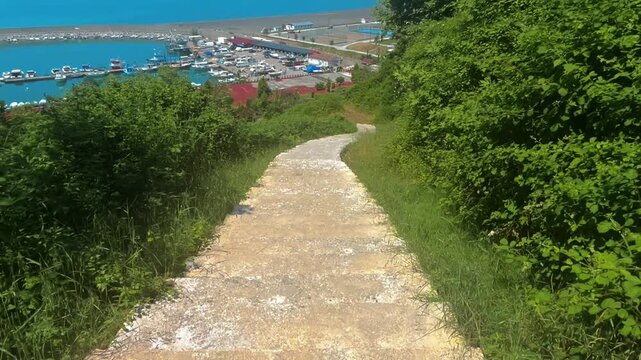 Cinematic point of view shot walking down outdoor stone stairs towards a beautiful sea harbor with boats and green trees on a sunny day.