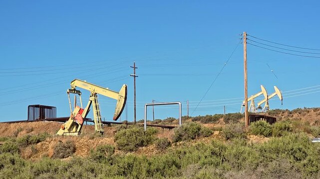 Oil pumpjacks stand on a dry hillside with power lines under a clear blue sky, Editorial video frame shows petroleum extraction equipment industrial pipes utility poles and visible machinery markings