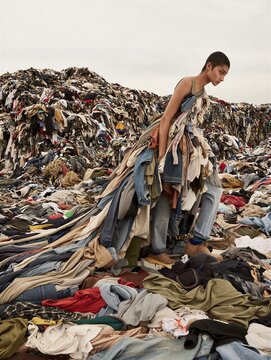 A fashion model stands amidst a vast pile of discarded clothes, wearing a dress crafted from various garments. Environmental awareness and sustainability theme.