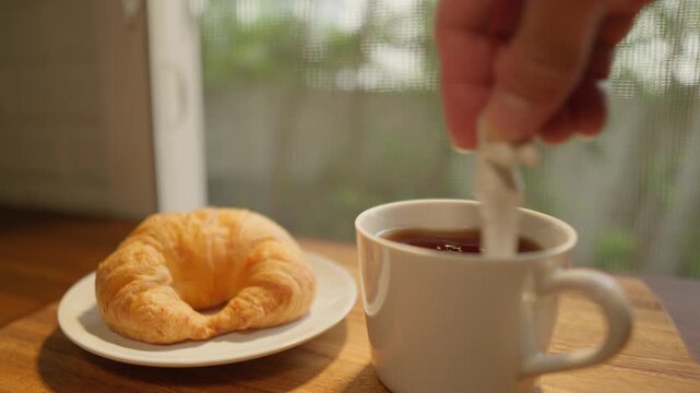 The camera pans from right to left, showing a wooden tray with a cup of freshly brewed coffee and a croissant. A spoon is then used to stir the coffee, creating a delicious breakfast amidst the soft m
