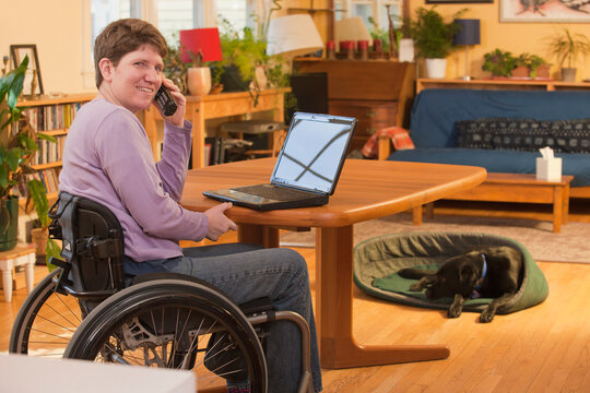 Disabled person in a wheelchair using a laptop at home and talking on the phone with her dog in the