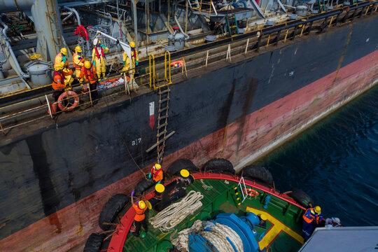 Aerial view of workers in safety gear on the deck of a large industrial ship alongside a tugboat with ropes and a ladder in Indonesia.