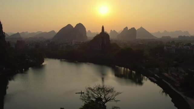 A tranquil sunset landscape featuring karst mountains, a calm lake, an arch bridge, and a pagoda silhouette, with reflections on the water.