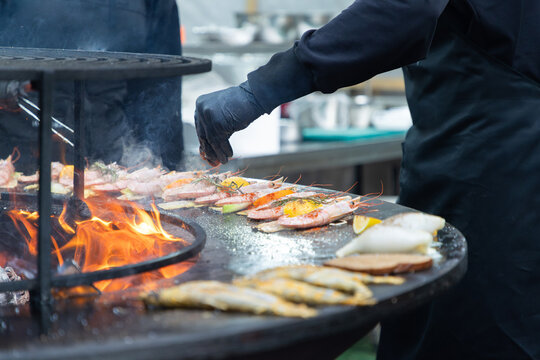 Unrecognizable chef wearing black gloves and apron grilling fresh prawns, fish, and squid on a large outdoor fire pit grill