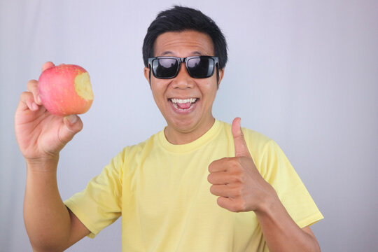 Happy Asian Man Wearing Sunglasses Holding Bitten Red Apple and giving thump up gesture