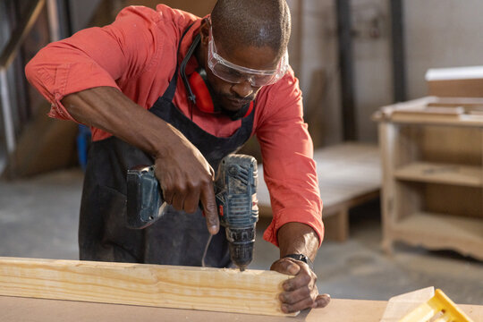 Cordless drill driving screw into wooden plank on workbench in workshop with scattered wood panels