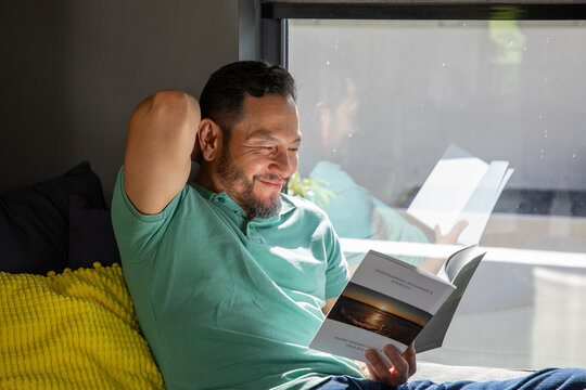 Mature Asian man reclining in mint polo on dark gray couch, reading book with window reflection