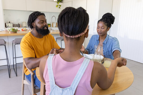 Parents and teenage daughter wearing pink top holding hands, bowing at kitchen table with pitcher