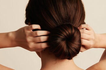 Fototapeta premium Woman with dark hair styles a neat bun. Her hands adjust the hairstyle carefully, creating a clean and elegant look. The studio shot focuses on her back and hands.