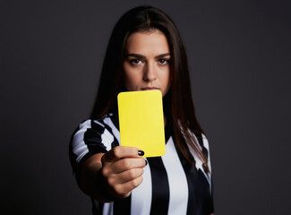 Obraz premium Young woman soccer referee in striped uniform shows yellow card. Serious female ref holds warning card for player foul in studio. Sport game official gives penalty.