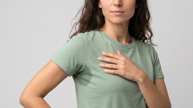 Woman wearing a sage green t-shirt with visible underarm sweat stains while standing in front of a neutral backdrop