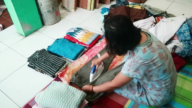A woman sits on the floor, folding colorful fabrics in a cluttered room with white tiles
