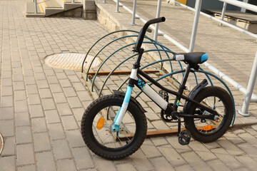 Obraz premium Small black and blue kids bicycle parked at metal rack on street sidewalk pavement. Child bike outdoor parking, urban transportation system infrastructure, active lifestyle concept