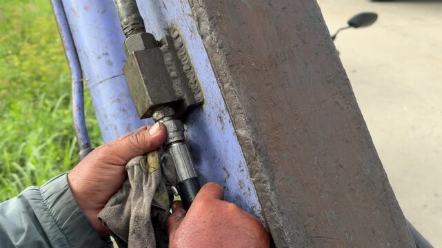 A worker secures a nut on a metal hydraulic component of an excavator with a wrench while performing adjustment beside grass vegetation and roadside ground. Industrial service.