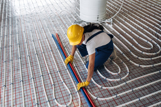 Construction worker in yellow hard hat and gloves installs heating pipes on the floor of a building under construction, with grid pattern visible on the surface
