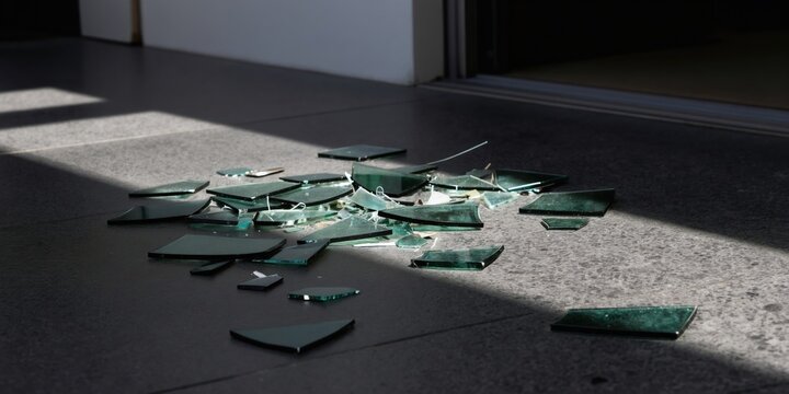 Scattered broken glass shards lie on a dark floor illuminated by sunlight. Sharp fragments create a hazardous mess indoors. Danger and damage are evident from the shattered material.