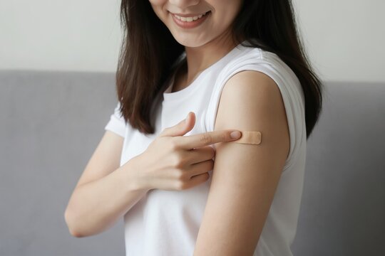 Young Asian woman shows arm with band aid after vaccine shot. She points to bandage, smiling after getting flu shot or corona jab. Health care concept.