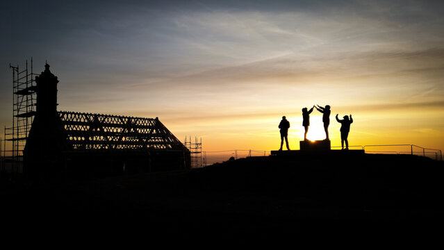 Aerial view of Chapelle Saint Michel de Brasparts under renovation with scaffolding and people silhouetted against a golden sunset sky in Saint-Rivoal, Bretagne, France.