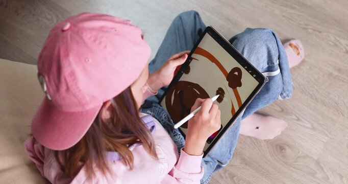 Child wearing pink cap drawing cute cartoon capybara on digital tablet screen with stylus pen. Creative educational graphic design software activity for early child development at home. Real indoor