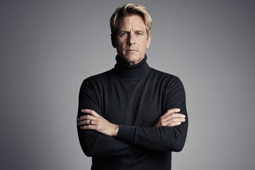 Obraz premium Mature blond man with arms crossed in a dark turtleneck sweater. His expression is calm and focused, standing against a plain grey background. He looks confident and serious.