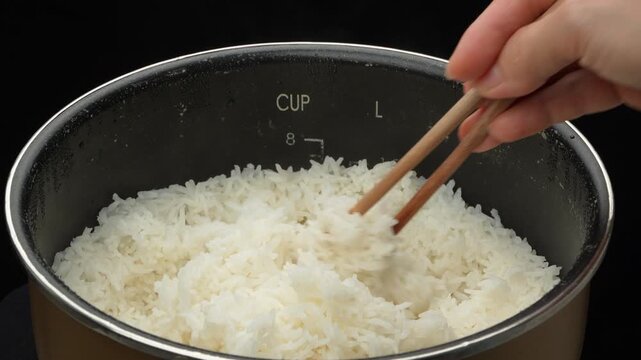 Close-up of freshly cooked ST25 white rice in an electric cooker pot with a hand using chopsticks to stir, Vietnam.
