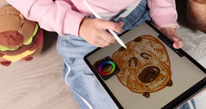 Child wearing pink cap drawing cute cartoon capybara on digital tablet screen with stylus pen. Creative educational graphic design software activity for early child development at home. Real indoor