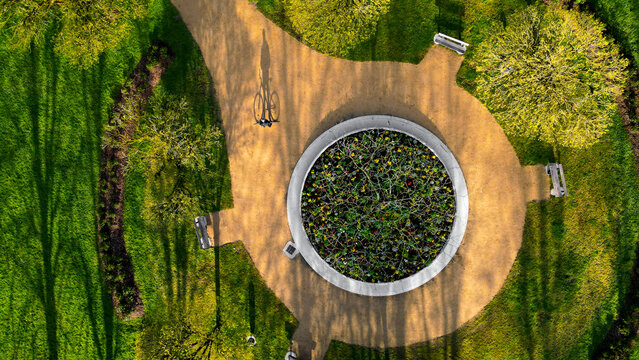 Aerial view of a circular flower bed surrounded by sandy paths and green grass with a person on a bicycle casting a long shadow in a public park in Morlaix, Brittany, France.