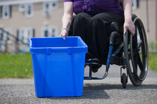 Woman with Spina Bifida in a wheelchair picking up a recycling bin at the street