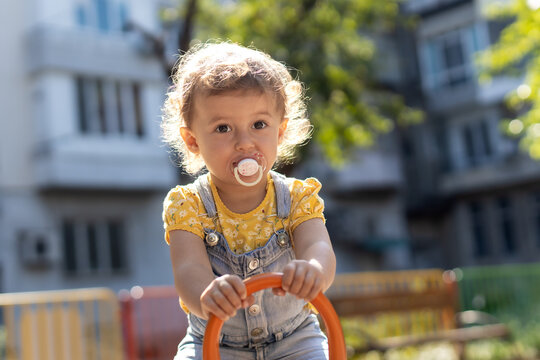 Adorable toddler girl with a pacifier and curly hair playing at an outdoor playground on a sunny day.