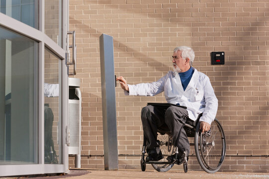 Doctor with muscular dystrophy in wheelchair at hospital entrance pressing knob for accessible door