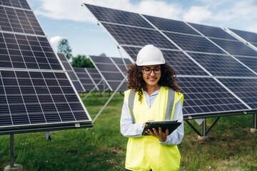 Obraz premium Female engineer wearing safety vest and helmet using tablet in solar farm with solar panels and green grass under blue sky on sunny day