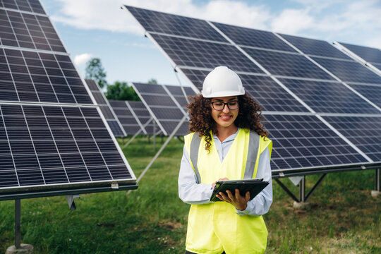 Female engineer wearing safety vest and helmet using tablet in solar farm with solar panels and green grass under blue sky on sunny day