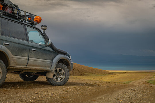 Side view of offroad SUV car with roof rack on dirt road by lake. Expedition 4x4 vehicle traveling through remote desert steppe under dramatic cloudy sky before storm.