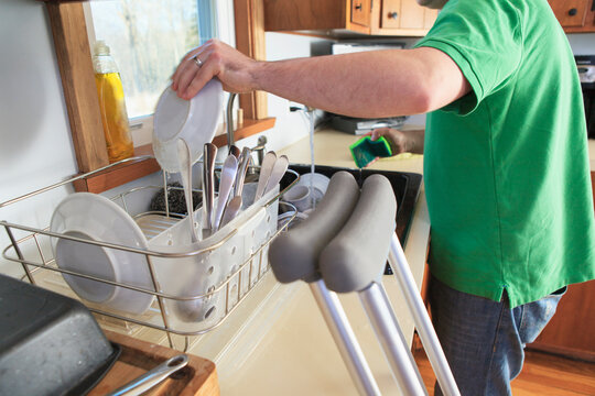 Man after anterior cruciate ligament (ACL) surgery with crutches washing dishes in the kitchen