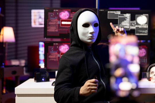 Hacker hidden in neon lit hideout films video ultimatum with smartphone, sending it to foreign governments, masked evil man threatens retaliation against foreign military targets.